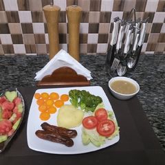 A picture of Mash Potatoes with some steamed veggies,sausages,fruit salad.