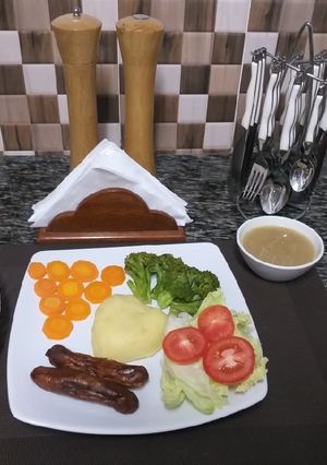 A picture of Mash Potatoes with some steamed veggies,sausages,fruit salad.