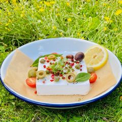 A picture of Baked Vegan Feta with olives, pink peppercorns, lemon and summer herbs and veggies 🌱.