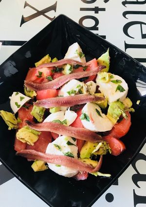 Una foto de Ensalada de tomate, mozzarella, aguacate y anchoas, con albahaca fresca y regada con Aceite oliva