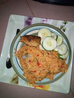 A picture of Jollof rice with chicken and sliced cucumber.