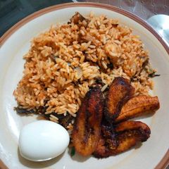 A picture of Dried catfish jollof rice with fried plantains and boiled egg.