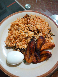 A picture of Dried catfish jollof rice with fried plantains and boiled egg.