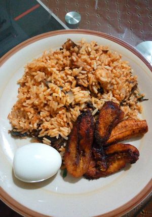 A picture of Dried catfish jollof rice with fried plantains and boiled egg.