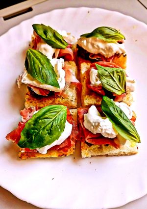 A picture of Pizza Crostini with Taggiasca Olive Spread, Stracciatella, Prosciutto, and Basil.